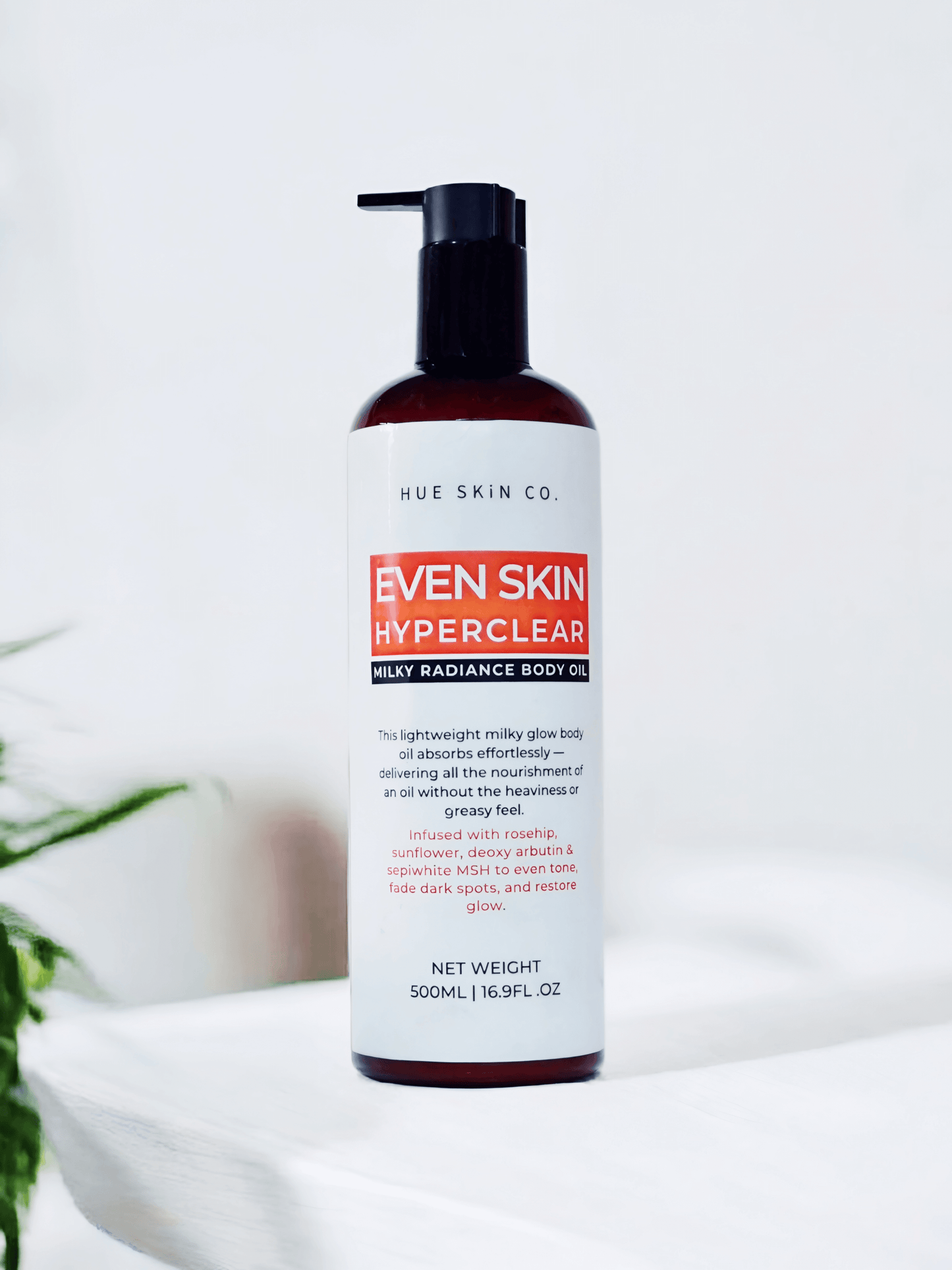 Even Skin HyperClear Milky Radiance Body Oil in a sleek bottle on a white background with a green plant.