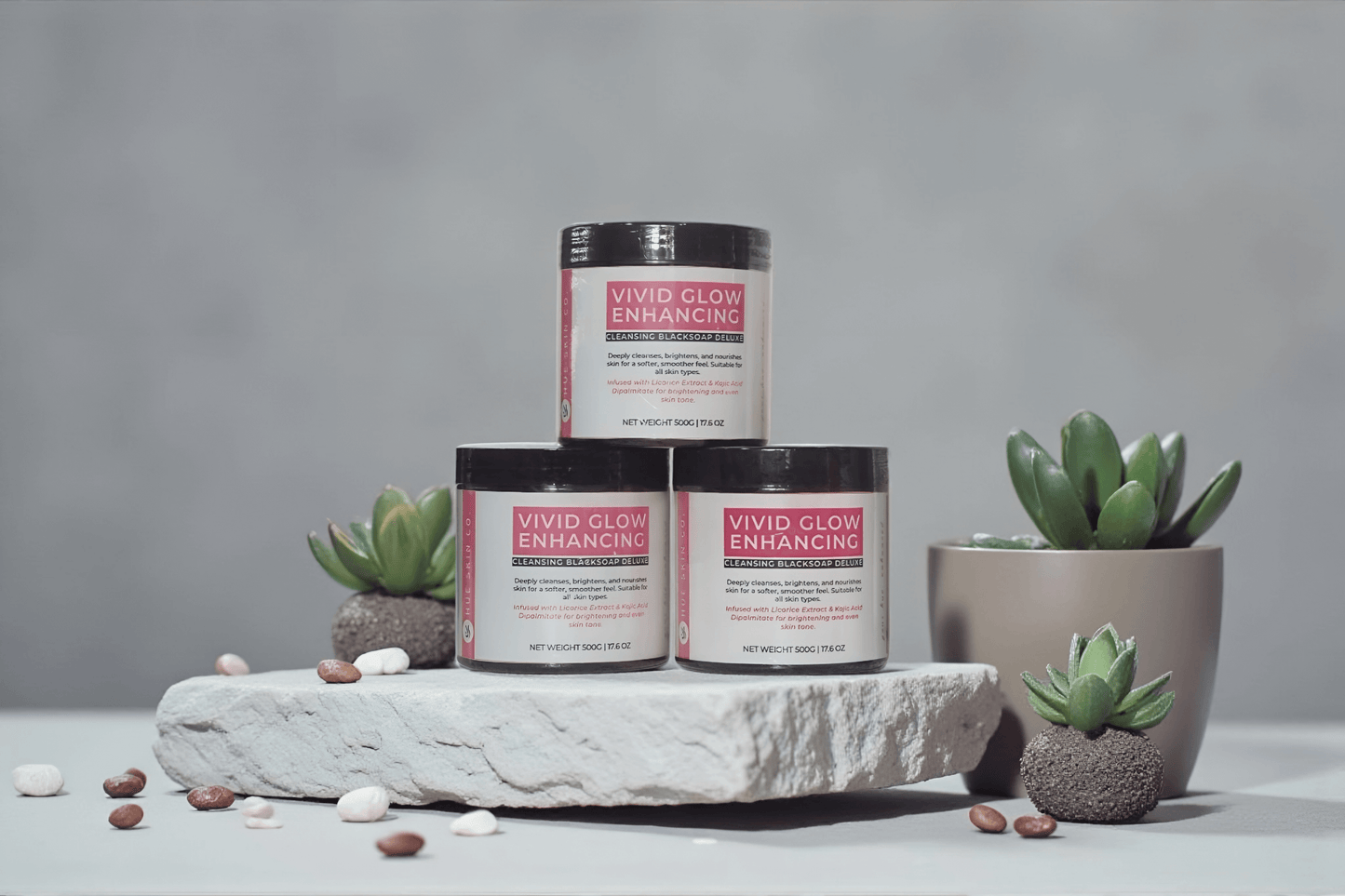 VIVID GLOW GLOW BLACK SOAP DELUXE OFFER - Three jars of enhancing soap on a stone display with decorative plants.